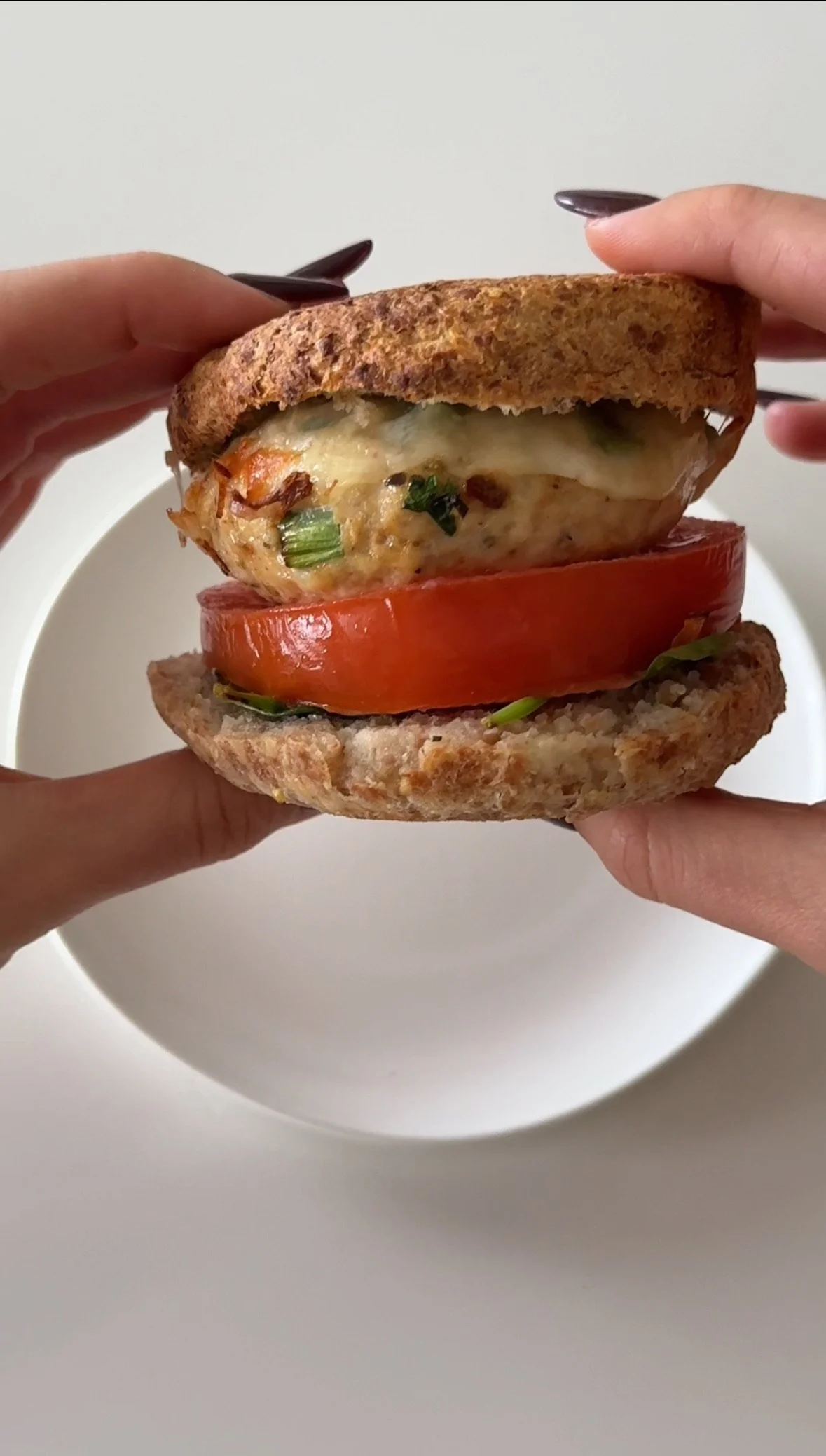 Close-up of a homemade chicken sandwich with melted cheese, tomato slices, and green herbs on whole grain buns, being held by two hands.