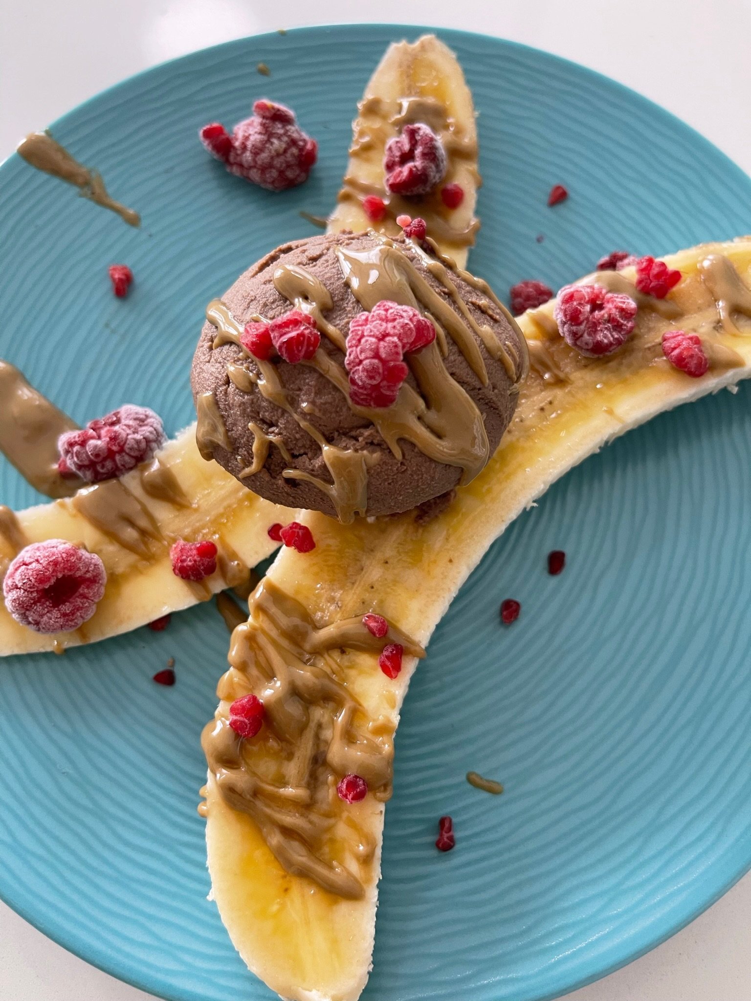 A sliced banana topped with caramel sauce and frozen raspberries on a blue plate with a scoop of chocolate ice cream drizzled with caramel sauce and raspberries.