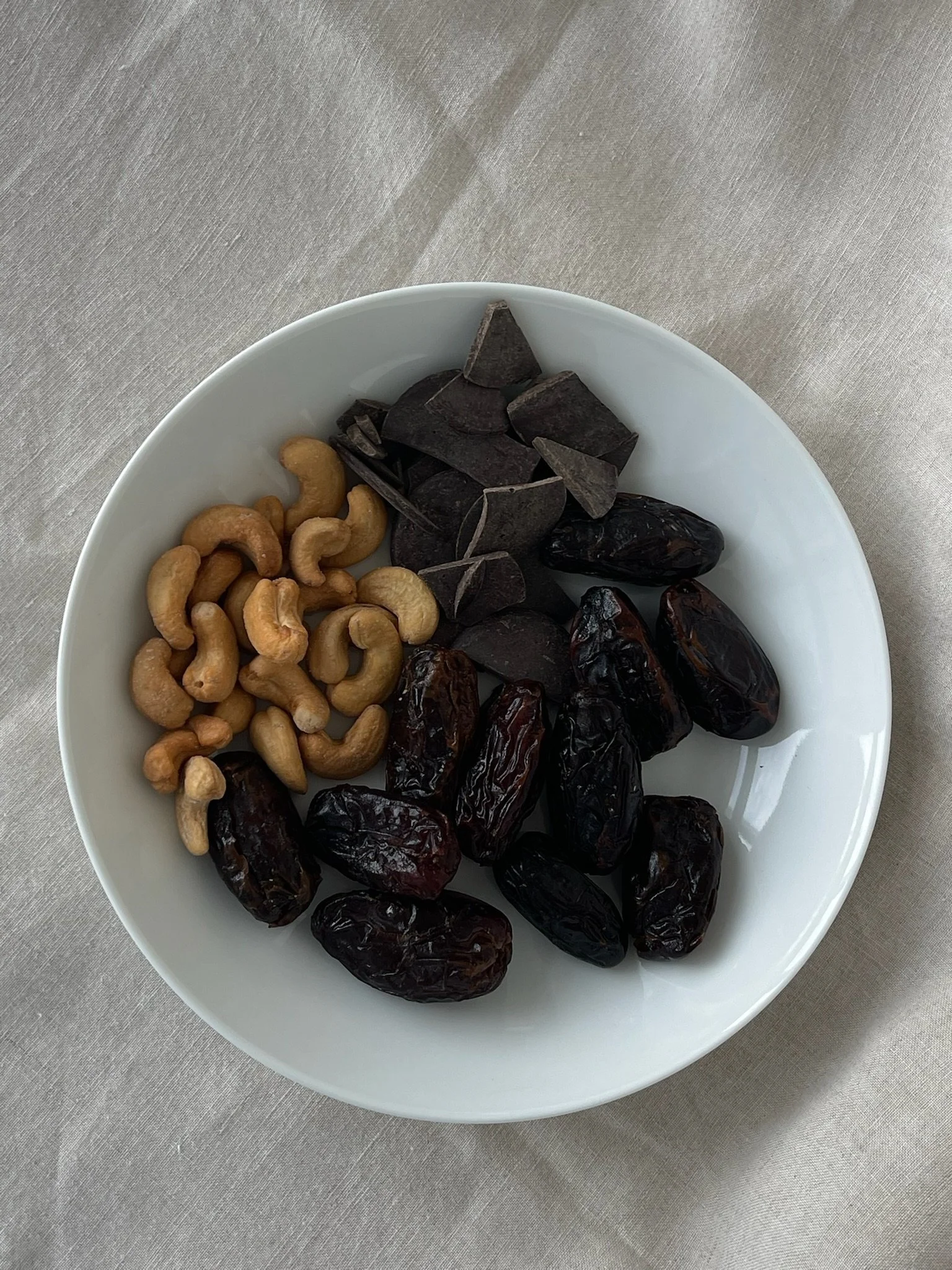 A white bowl filled with cashews, black chocolate chunks, dates, and black dried figs, placed on a beige cloth surface.