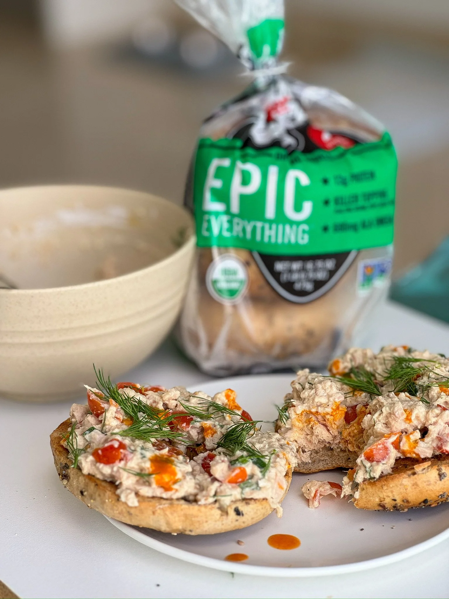 Open-faced bagel sandwiches topped with tuna salad, garnished with dill and hot sauce, on a white plate.