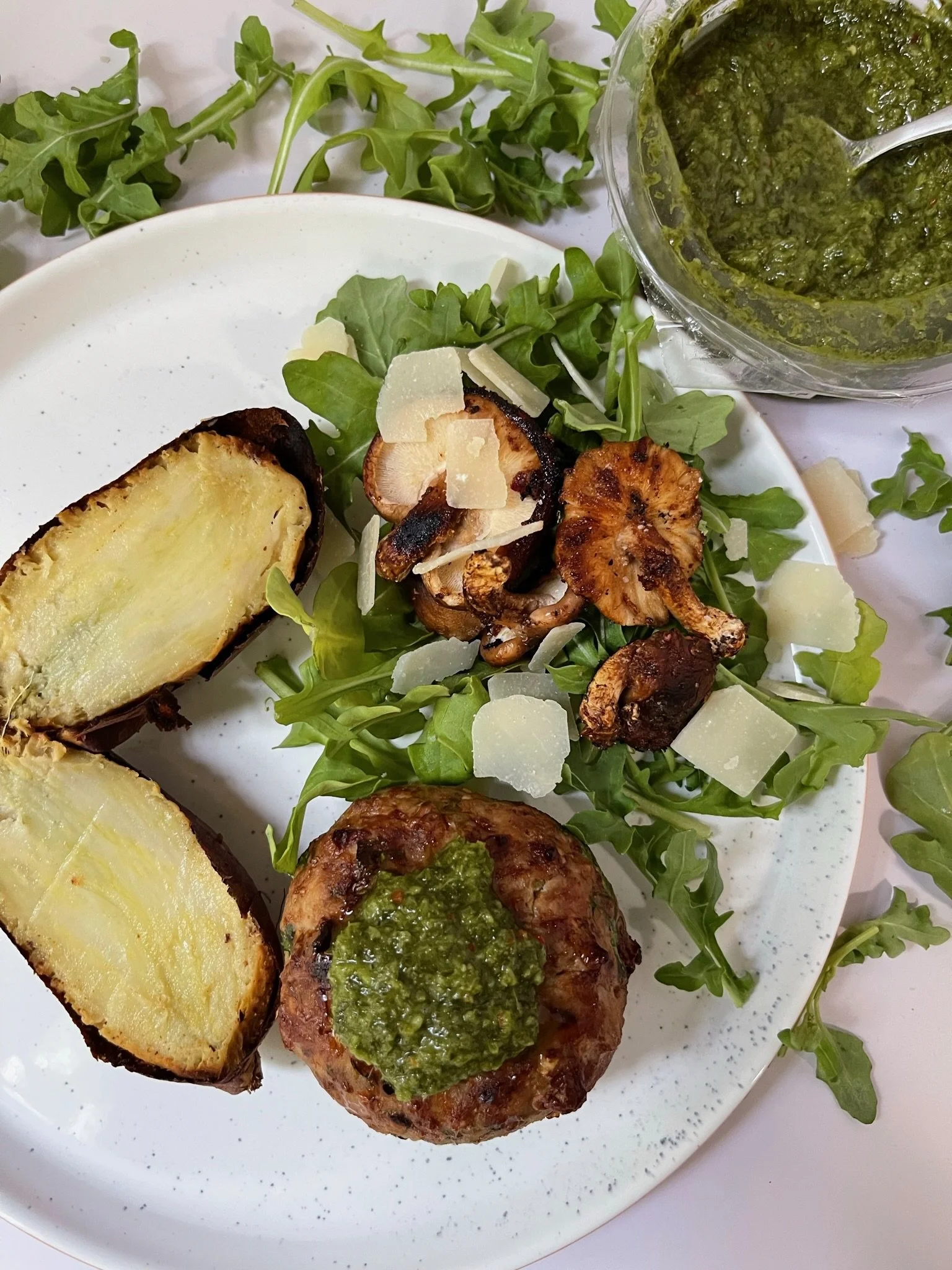 Plate with salad greens, roasted eggplant slices, grilled mushrooms, shaved cheese, and a meat patty topped with green pesto, served with a side of chimichurri sauce in a container.