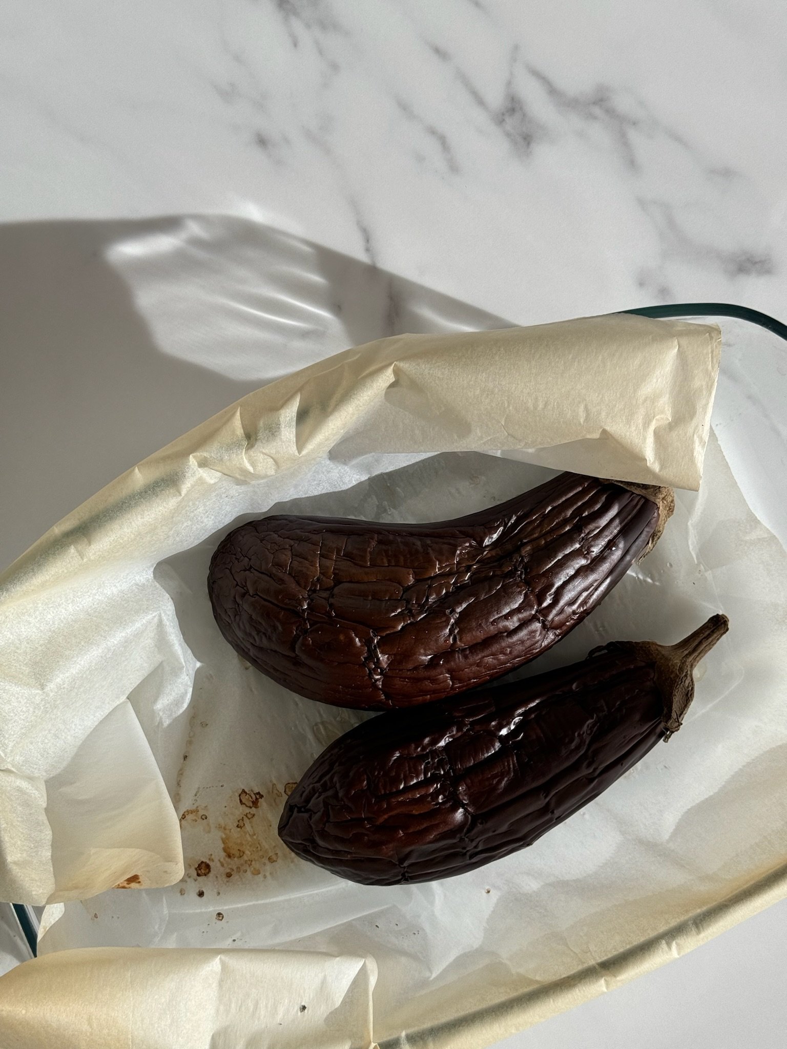 Two roasted eggplants on white parchment paper inside a glass baking dish, with a marble countertop in the background.