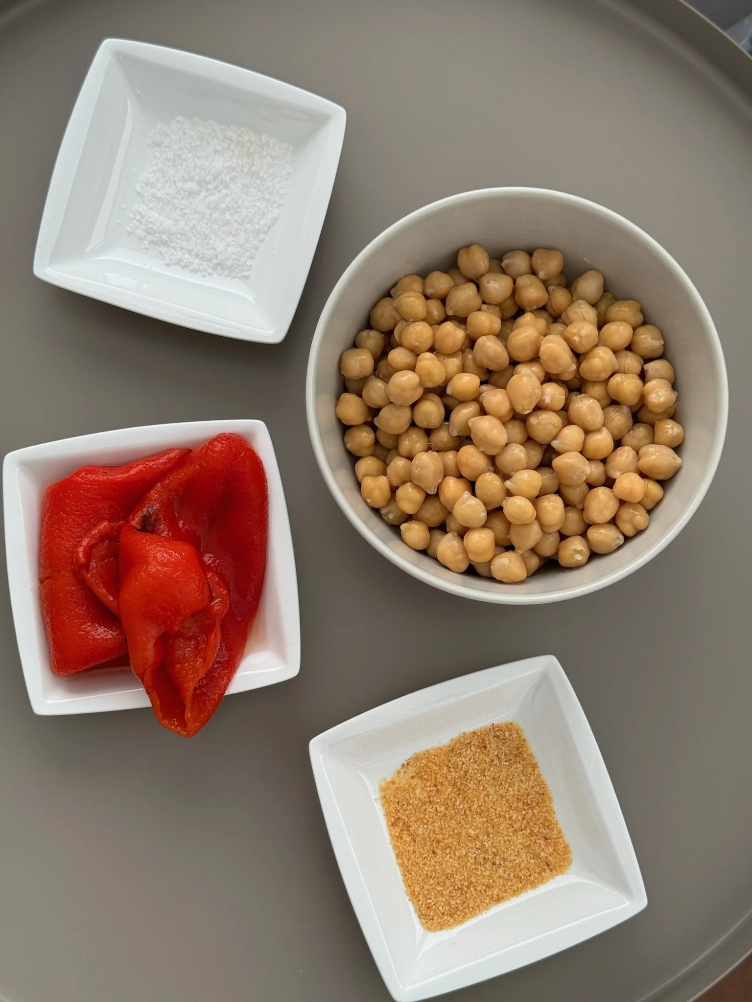 A white bowl filled with cooked chickpeas, a small square dish with red roasted peppers, a small square dish with salt, and a small square dish with seasoning or spice, all placed on a gray surface.