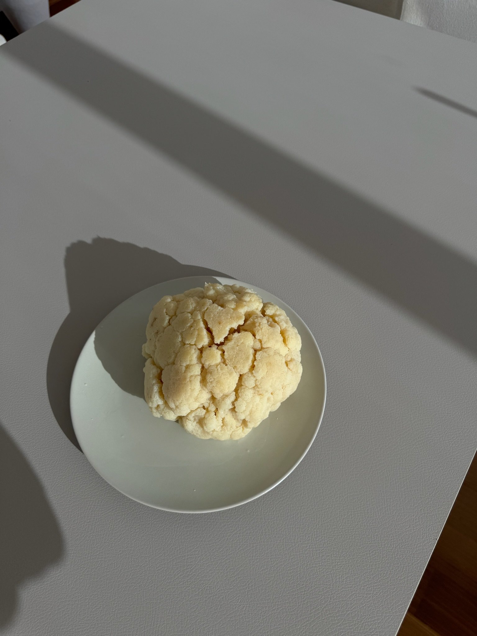 A head of cauliflower on a white plate, placed on a white surface with shadows cast across it.