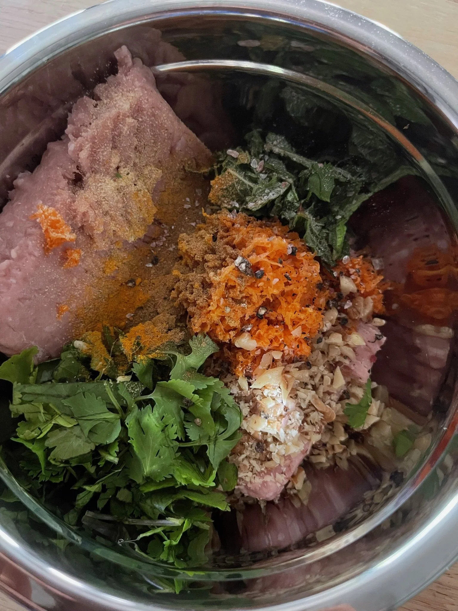 A metal mixing bowl containing raw meat, chopped cilantro, shredded carrots, and various spices and seasonings.