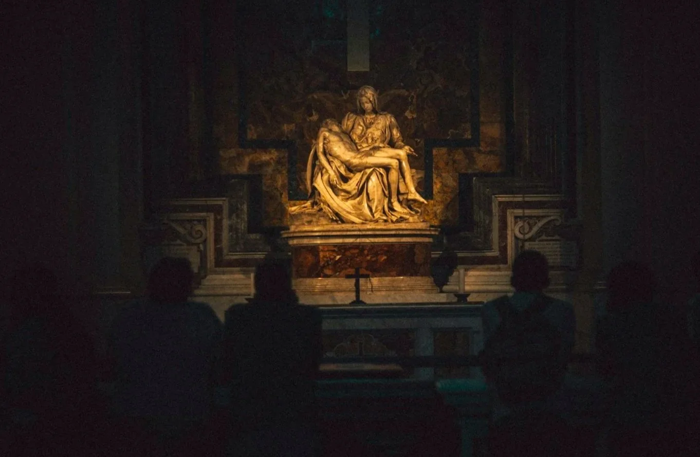 Réplica de la escultura de La Pietà en una capilla oscura, iluminada cuidadosamente, con varias personas contemplándola.