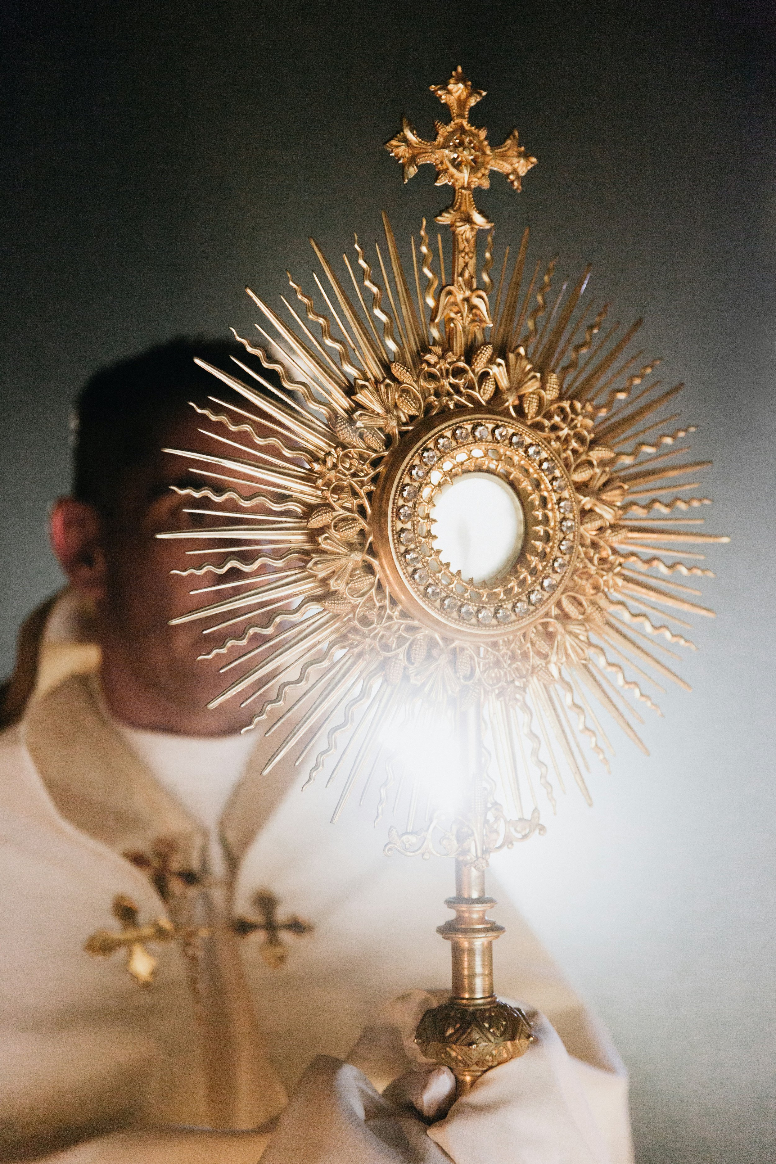 Un sacerdote sostiene un ostensorio dorado con radiantes detalles y piedras preciosas, utilizado en ceremonias religiosas católicas.