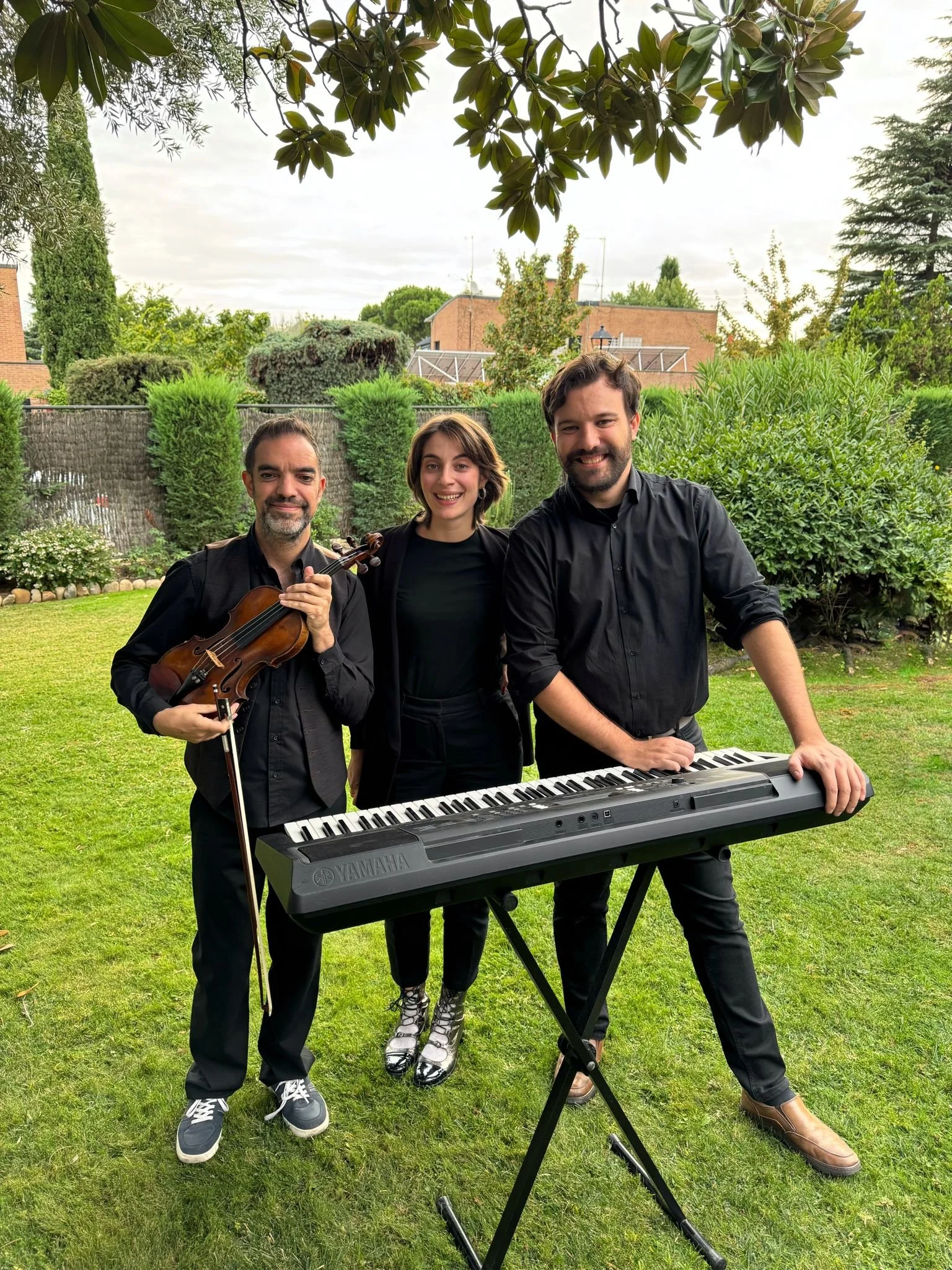Tres músicos con instrumentos en un jardín, uno con violín, otro con teclado y una mujer en medio, sonriendo.