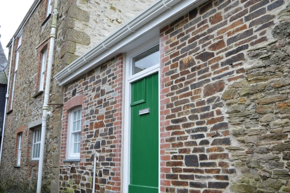 Close-up of a brick house with three white-framed windows, a green door, and a white drainpipe on the exterior wall.