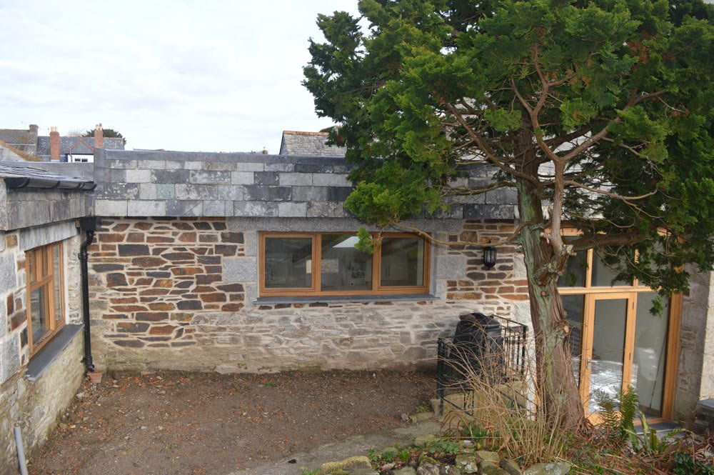 A stone building with a flat roof, three windows with wooden frames, a glass door, a large tree in front, and an outdoor area with dirt ground and some plants.