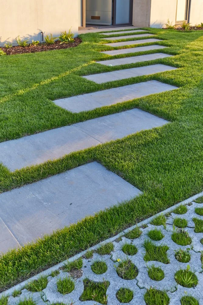 Concrete stepping stones arranged in a pathway on a lush green lawn, with a section of patterned concrete tiles with grass growing through circular holes in the foreground, leading to a house entrance.