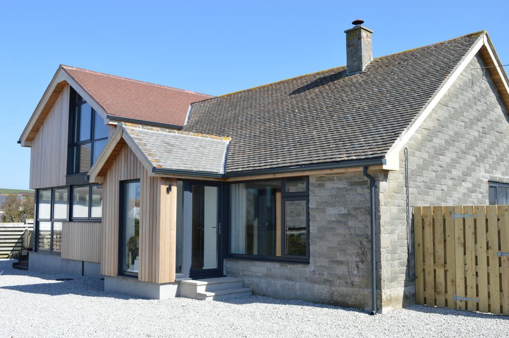 A modern two-story house with a mix of gray brick and light wooden siding, large windows, and a pitched roof with gray shingles.