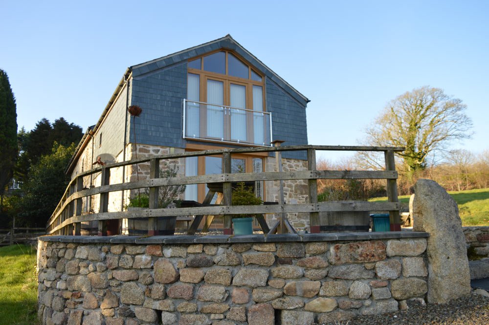 A two-story stone and wood house with large windows and a balcony, surrounded by a wooden fence and situated on a stone foundation, with trees in the background.