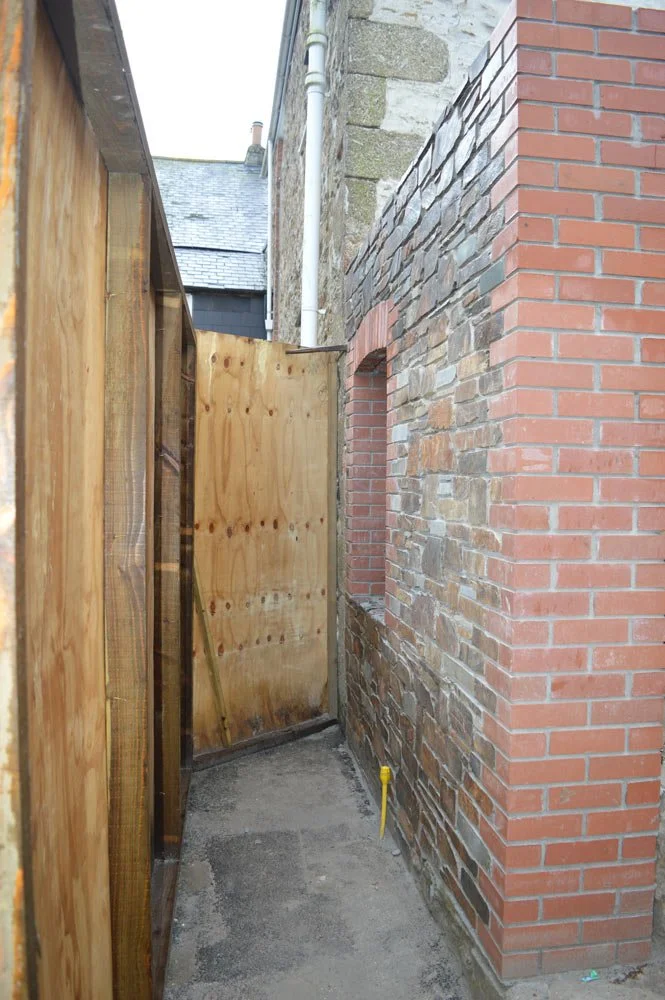 A narrow alleyway with a brick building on the right and a wooden construction on the left, featuring a small window opening and a concrete floor.