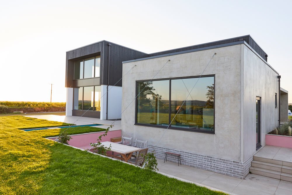 Modern residential house with large windows, a patio table and chairs, and a green lawn during sunset.