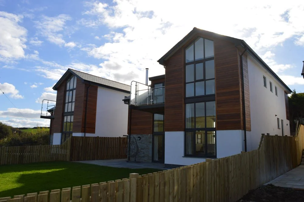 Modern two-story house with large glass windows, wooden siding, white walls, a small balcony, a fenced backyard, and a sunny sky.
