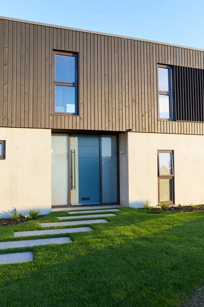 Modern two-story house with a green front lawn, a stone walkway leading to the front door, and a combination of wood and concrete exterior walls.