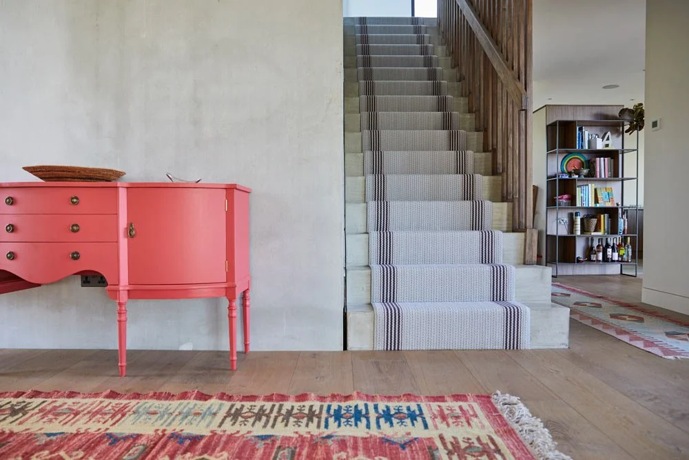 Interior of a home showing a pink sideboard, a staircase with a striped runner, and a black bookshelf with various decorative items.