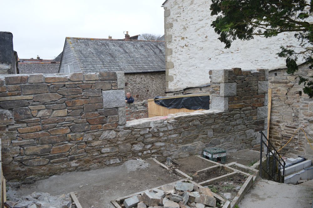 Construction site with stone walls and stairs, partially built patio, and construction materials, with neighboring brick buildings and trees in the background.