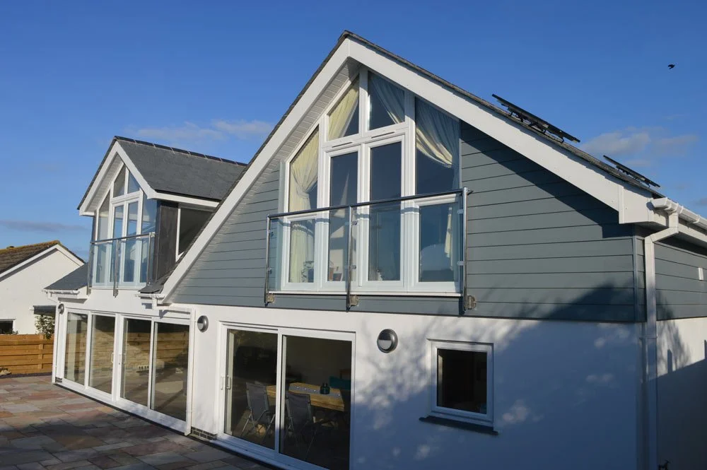 A modern two-story house with large windows, a balcony, and solar panels on the roof, under a clear blue sky.