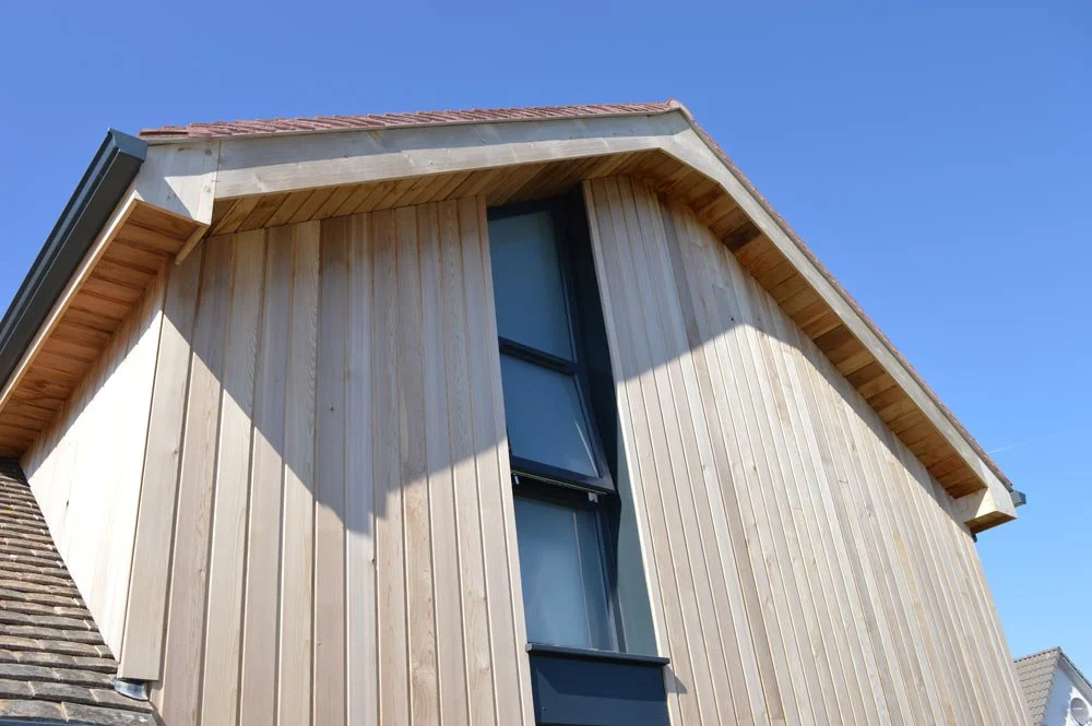 Close-up of a modern house exterior with vertical light wood siding, a large vertical window, and a sloped roof against a blue sky.