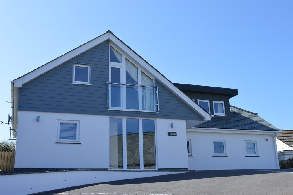A modern two-story house painted in white and gray, with large windows and a sloped roof, situated under a clear blue sky.