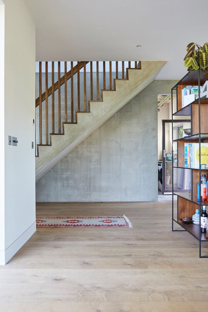 Interior view of a modern home with a staircase, wooden railings, a bookshelf, and a colorful rug on wooden flooring.