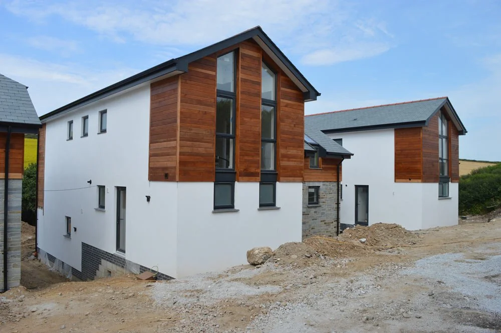 Modern new residential building with white walls, wooden accents, and multiple windows, situated on a construction site with dirt ground and a cloudy sky.