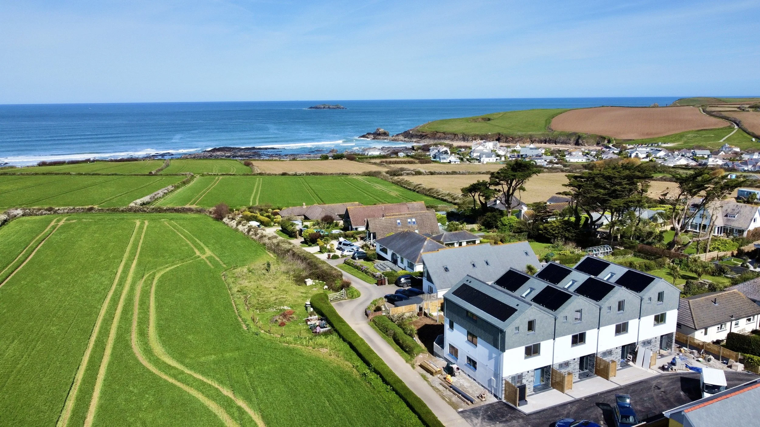 Aerial view of a coastal neighborhood with modern houses, green and brown farmland, and a large body of water with small islands in the distance.