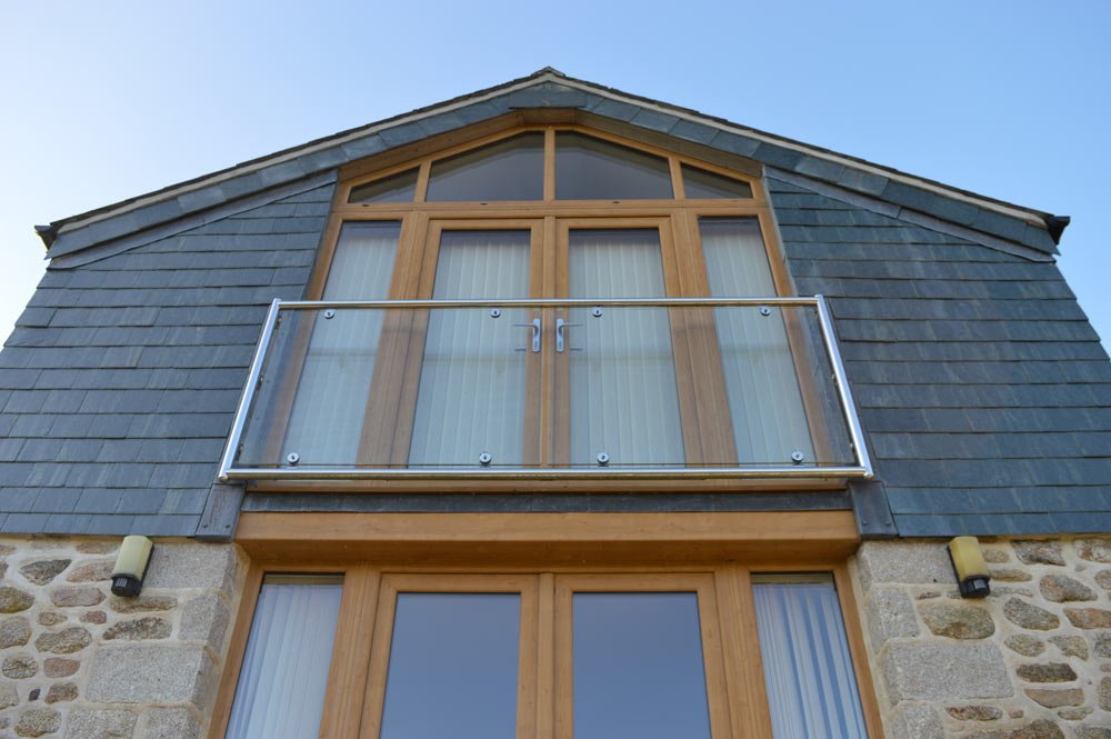 Exterior view of a house with a balcony, glass railing, and large windows, featuring stone and wood siding under a clear blue sky.