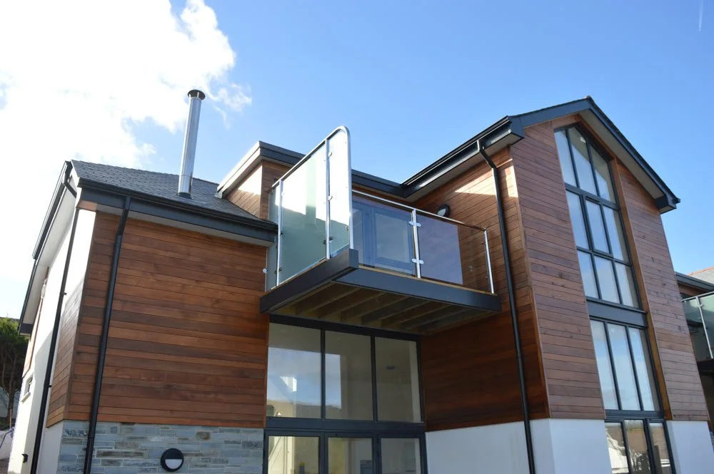Modern multi-story house with wooden siding, large windows, and a small balcony with glass railing, under a blue sky with clouds.