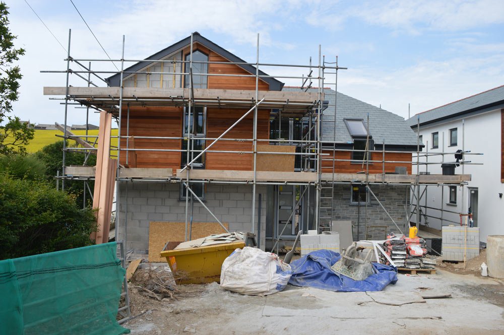 Construction site of a two-story house with scaffolding surrounding it, partly finished with wood and brick walls, and construction materials and tools on the ground.