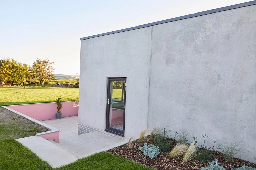 Exterior of a modern house with a pink wall, glass door, and landscaped garden with grass, plants, and trees.