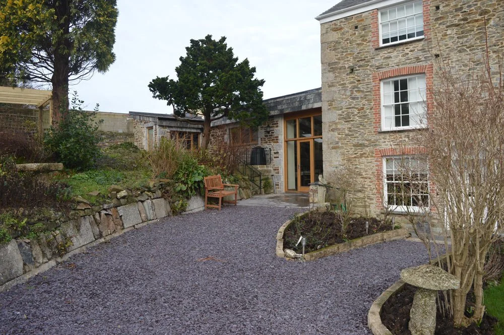 Backyard of a stone house with a gravel patio, wooden bench, plants, and trees.