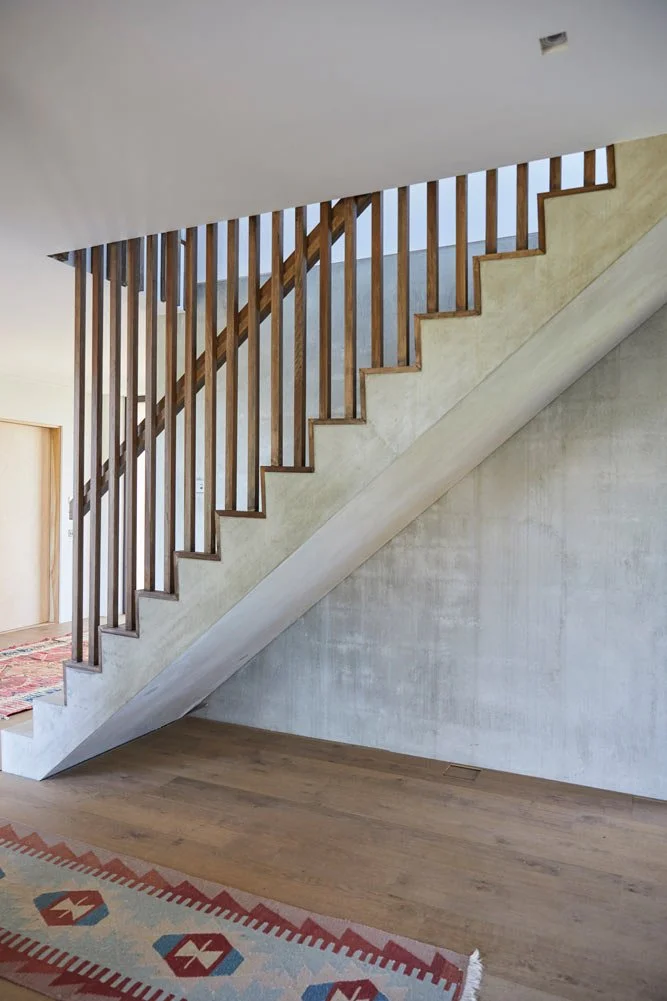 Interior staircase with wooden handrails and vertical balusters, concrete stairs, and hardwood flooring. A patterned rug is partially visible at the bottom.