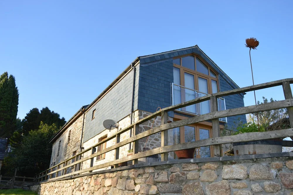 A house with stone and wood exterior, large glass windows, and a sloped roof, situated on a stone foundation with a wooden fence in front.