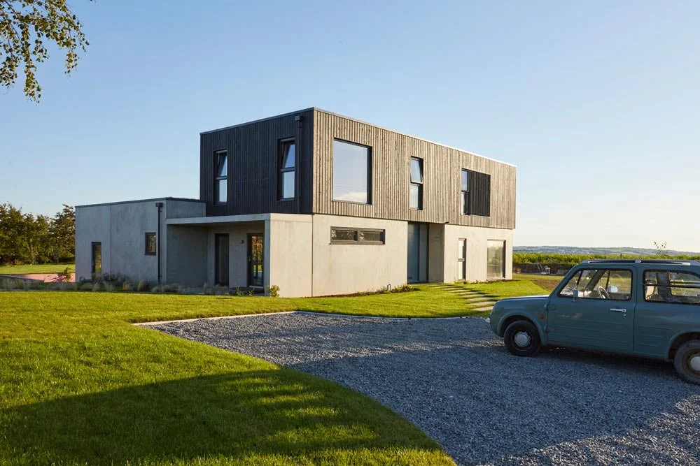 Modern two-story house with concrete and wood exterior, green lawn, gravel driveway, and vintage blue car parked outside.
