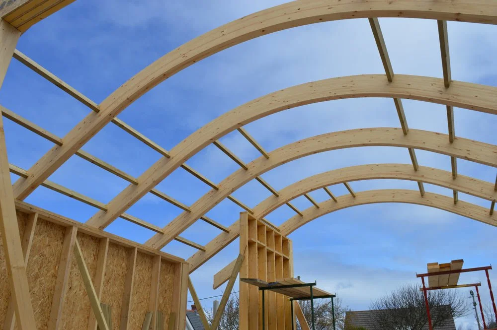 The image shows the wooden framework of a building under construction with curved arches made of timber, against a blue sky with some clouds.