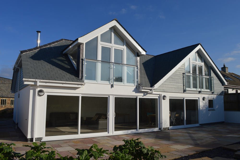 Modern two-story house with large glass sliding doors and windows, small balconies on the upper floor, and a stone-paved patio in front.
