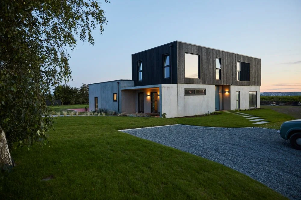 Modern two-story house with a black upper floor and white lower floor, surrounded by green grass and trees, with a gravel driveway at sunset.
