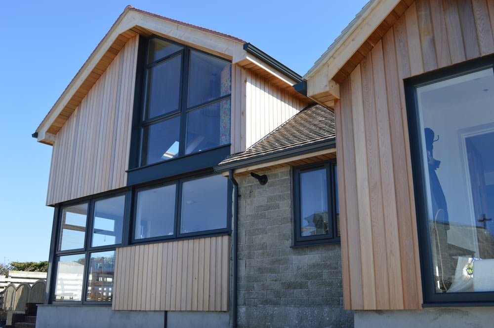 A modern house with large glass windows, wood siding, and a stone and shingle exterior under a clear blue sky.