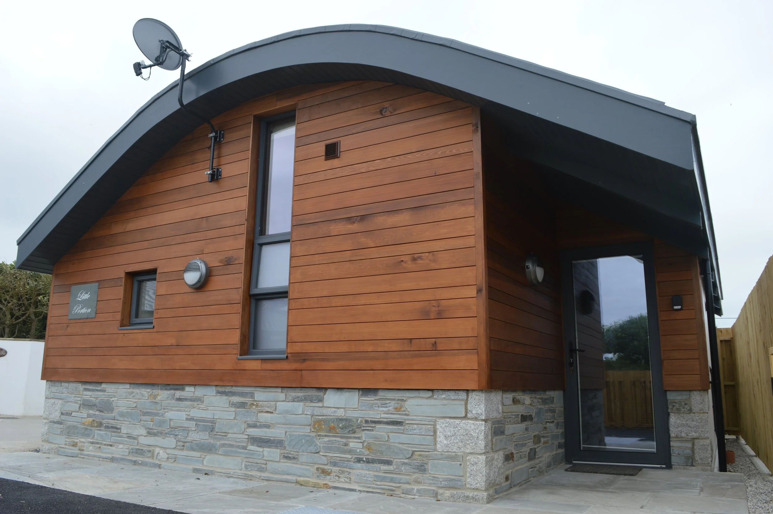 Front view of a modern house with wooden siding, a stone foundation, and angular roof, featuring a satellite dish mounted on the roof.