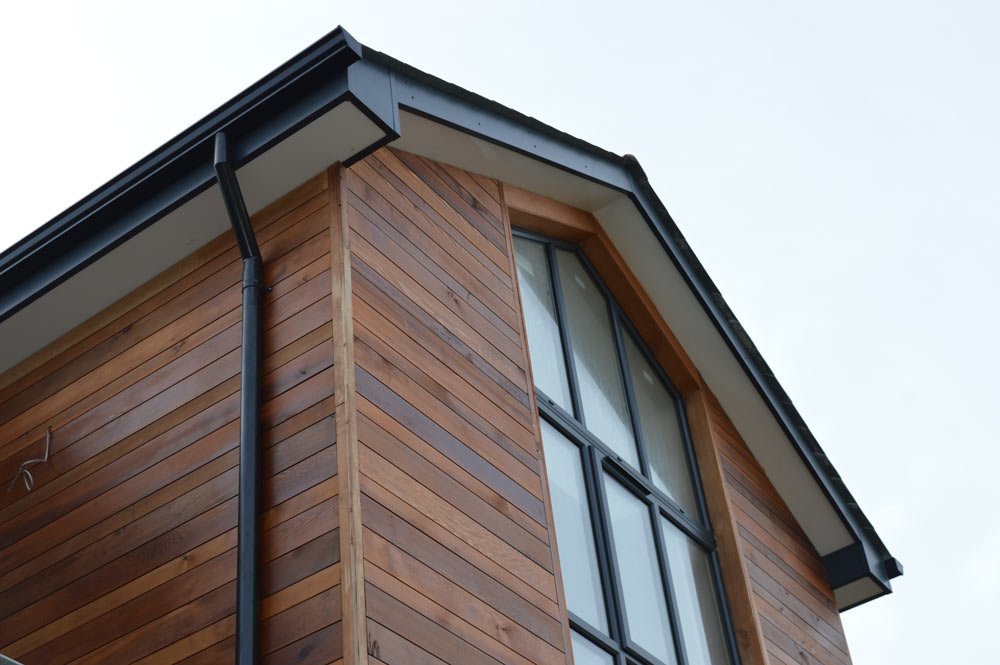 Close-up of a modern house corner with wooden siding and large glass windows, black rain gutter, and overcast sky.