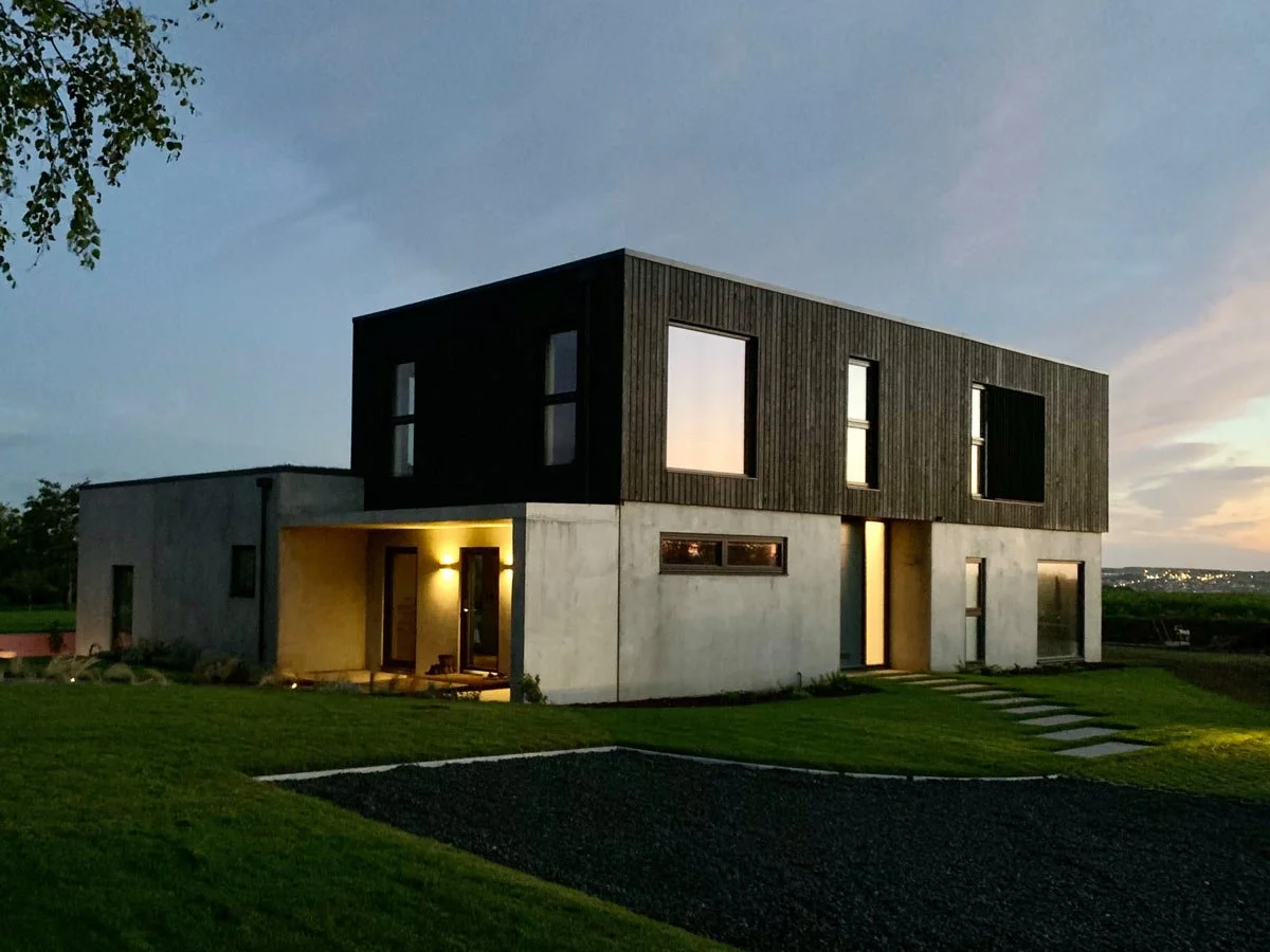 Modern two-story house with concrete and black wood exterior, illuminated entryway, surrounded by green lawn, at dusk.