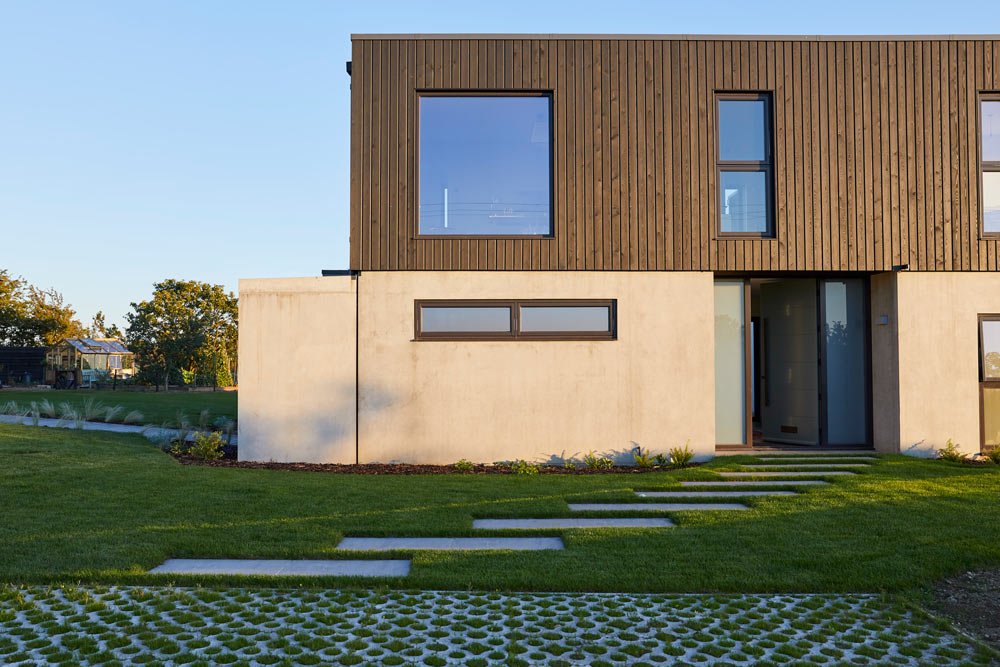 Modern two-story house with a combination of brown wood and white concrete exterior, large windows, and a pathway leading to the front door, set against a background of green lawn and trees.