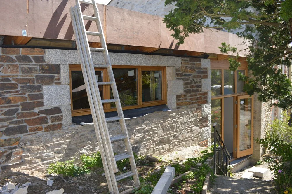 Construction site of a stone house with a ladder leaning against the wall, and partially installed windows and door.