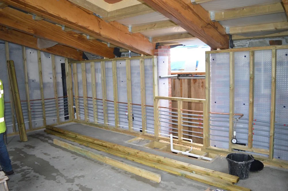 Construction site showing interior framing with electrical and plumbing rough-ins, wooden beams, and insulated wall panels.