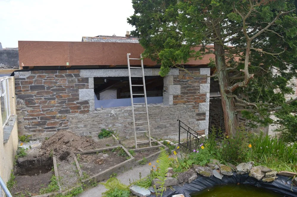 Construction site with a partially built stone and brick wall, a wooden panel on top, a ladder leaning against the wall, and a tree on the right side with garden plants and flowers in front.