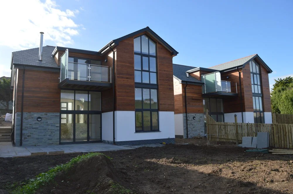 New modern multi-family residential building with large windows, wooden exterior, and small balconies, located in a developing neighborhood.