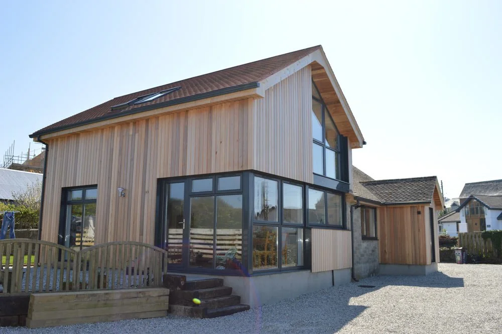 Modern two-story house with wooden siding, large windows, and a sloped roof, set in a residential neighborhood.